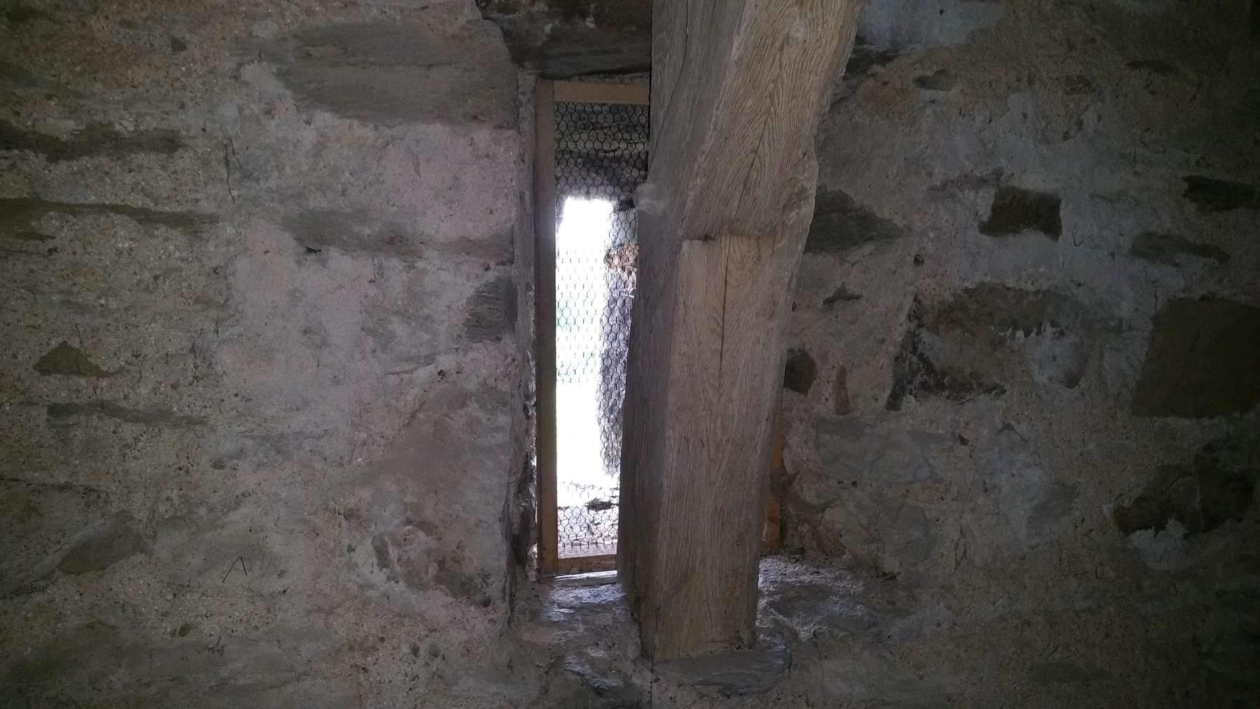 Schmales vergittertes Fenster im Bruchsteinmauerwerk des Turms mit einem Holzbalken davor
