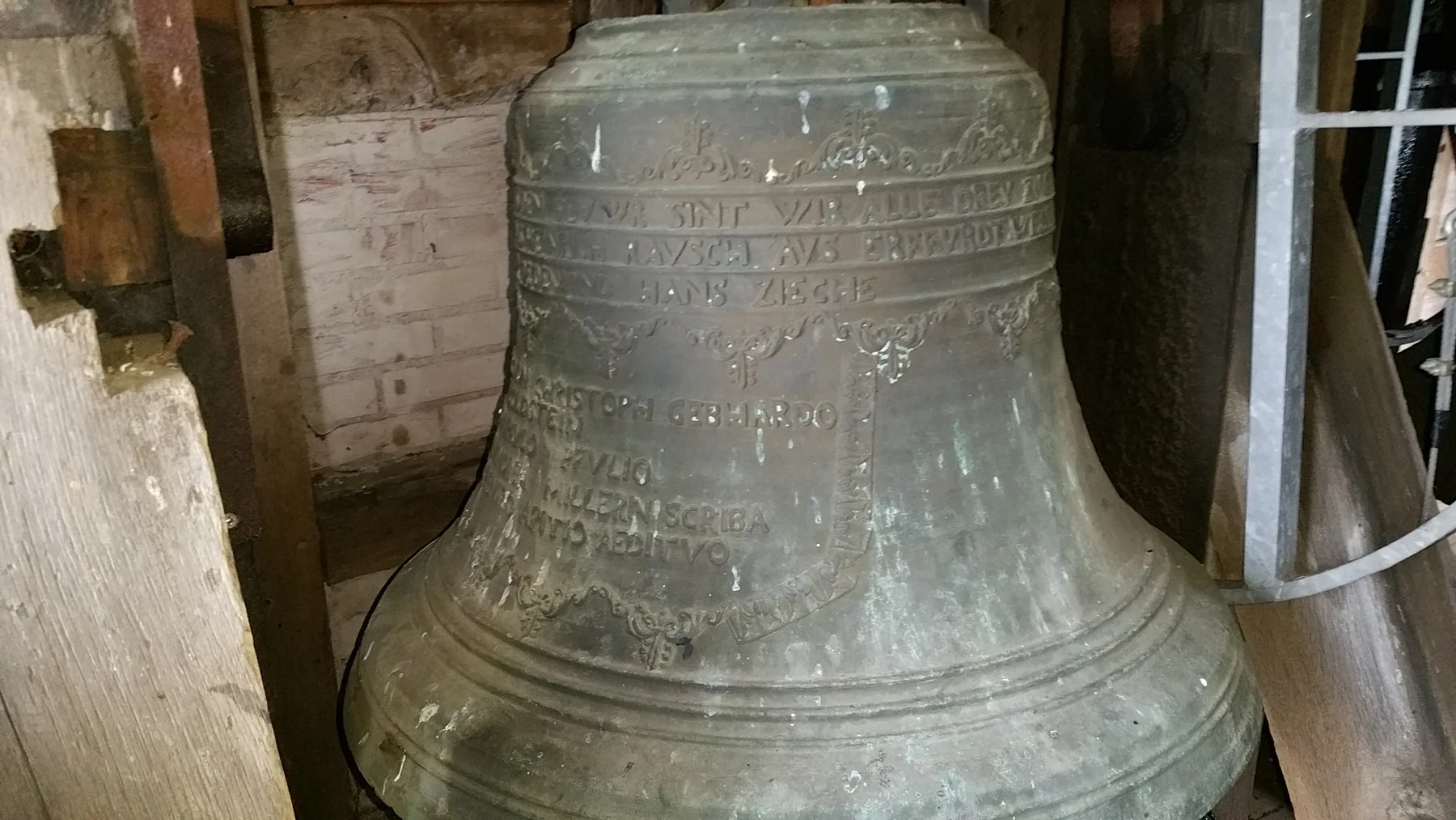 Nahaufnahme einer Glocke mit umlaufender Inschrift und Ornamenten zwischen den Holzbalken des Glockenturms